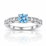 A silver-toned diamond ring featuring a round-cut light blue center stone and small clear accent stones set in square-shaped bands, shown on a white background with a soft reflection below.