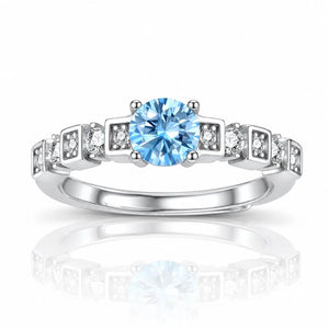 A silver-toned diamond ring featuring a round-cut light blue center stone and small clear accent stones set in square-shaped bands, shown on a white background with a soft reflection below.