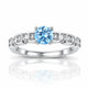 A silver-toned diamond ring featuring a round-cut light blue center stone and small clear accent stones set in square-shaped bands, shown on a white background with a soft reflection below.