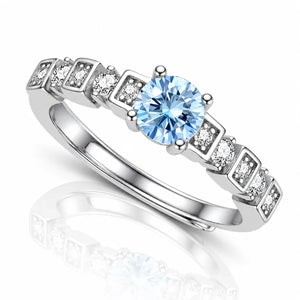 A silver-toned ring featuring a round light blue center stone and a row of square-cut clear accent stones on a white background with a soft reflection.