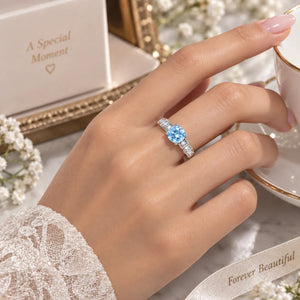 A close-up lifestyle shot of a hand wearing a silver ring with a round light blue gemstone, holding a white teacup on a saucer. In the background, there is a white jewelry box with the text 'A Special Moment' and a white ribbon with 'Forever Beautiful'.