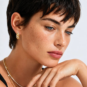 Close-up of a woman with short dark hair wearing a small gold hoop earring, layered gold and pearl necklaces, and a black top against a light gray background.