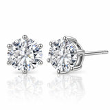 A pair of round-cut diamond stud earrings in a silver four-prong setting, with one earring facing forward and the other angled to show the post and back, on a reflective white surface.