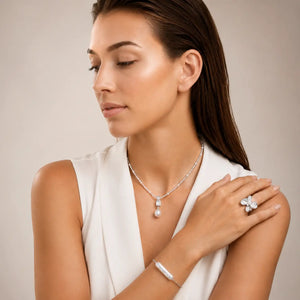 A woman wearing a silver-toned jewelry set including a delicate pendant necklace, a matching bracelet, and a statement ring, styled with a white sleeveless top against a neutral beige background.