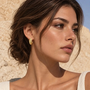 A close-up of a woman wearing a small gold crescent-shaped hoop earring, set against a warm beige stone wall background.