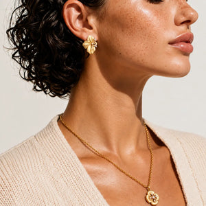 A close-up lifestyle shot of a model wearing a matching gold jewelry set, featuring a textured flower-shaped stud earring and a delicate chain necklace with a matching flower pendant, styled with a cream-colored knit cardigan.