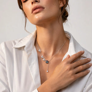 A close-up of a woman wearing a delicate silver necklace with a butterfly and flower pendant, styled with a white collared shirt against a light gray background.