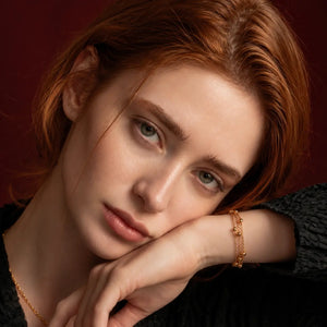 A close-up portrait of a woman with red hair resting her head on her hand, wearing a delicate gold chain necklace and a gold bracelet against a dark red background.