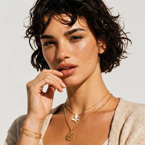 A close-up lifestyle shot of a woman with short dark curly hair wearing layered gold necklaces, including a white butterfly pendant and a coin pendant, and a gold bracelet, against a clean off-white background.