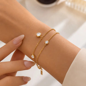A close-up of a person's wrist wearing two delicate gold chain bracelets, each adorned with a round clear stone, against a softly blurred light background.
