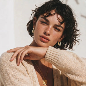 Woman wearing a beige sweater and gold jewelry against a neutral background