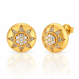 A pair of round yellow gold stud earrings with a starburst design paved with clear gemstones, shown on a reflective white surface.