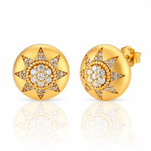 A pair of round yellow gold stud earrings with a starburst design paved with clear gemstones, shown on a reflective white surface.