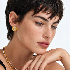 A close-up portrait of a model with short dark hair wearing a gold floral stud earring, layered gold and pearl necklaces, and a black top against a clean white background.