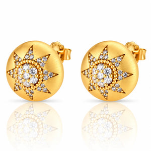 A pair of round gold stud earrings with a starburst design set with clear gemstones, shown on a white background with a soft reflection below.