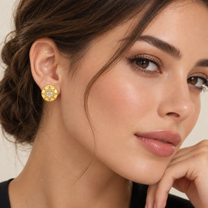 A close-up of a woman wearing a gold-toned geometric stud earring with clear stones, shown against a soft beige background.