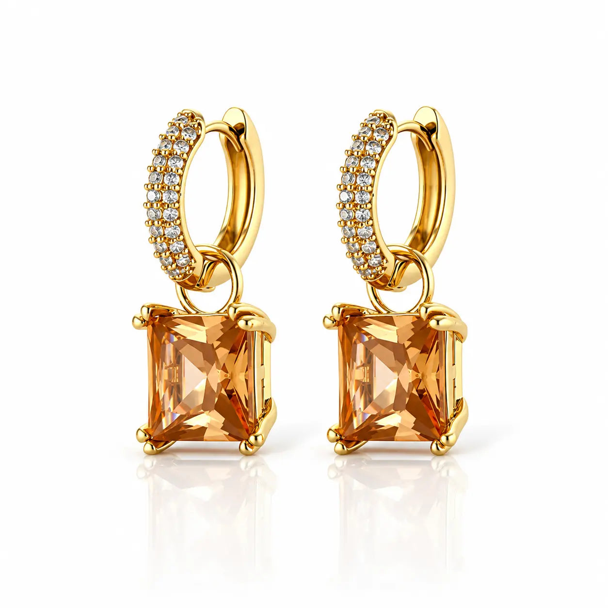 A pair of gold huggie hoop earrings with a dangling square-shaped orange gemstone and a row of small clear stones on the hoops, shown on a white background with a soft reflection.