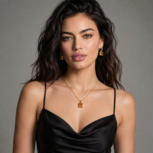 A woman with long dark hair models a gold necklace and matching drop earrings with square-cut clear stones, wearing a black satin camisole against a neutral gray background.