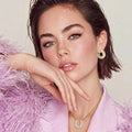 Woman wearing gold hoop earrings and a necklace, sitting on a pink fluffy surface.