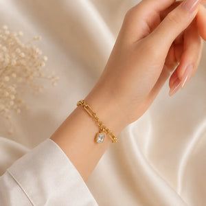 A delicate gold chain bracelet with a small clear rectangular gemstone charm, worn on a woman's wrist against a soft beige satin background.