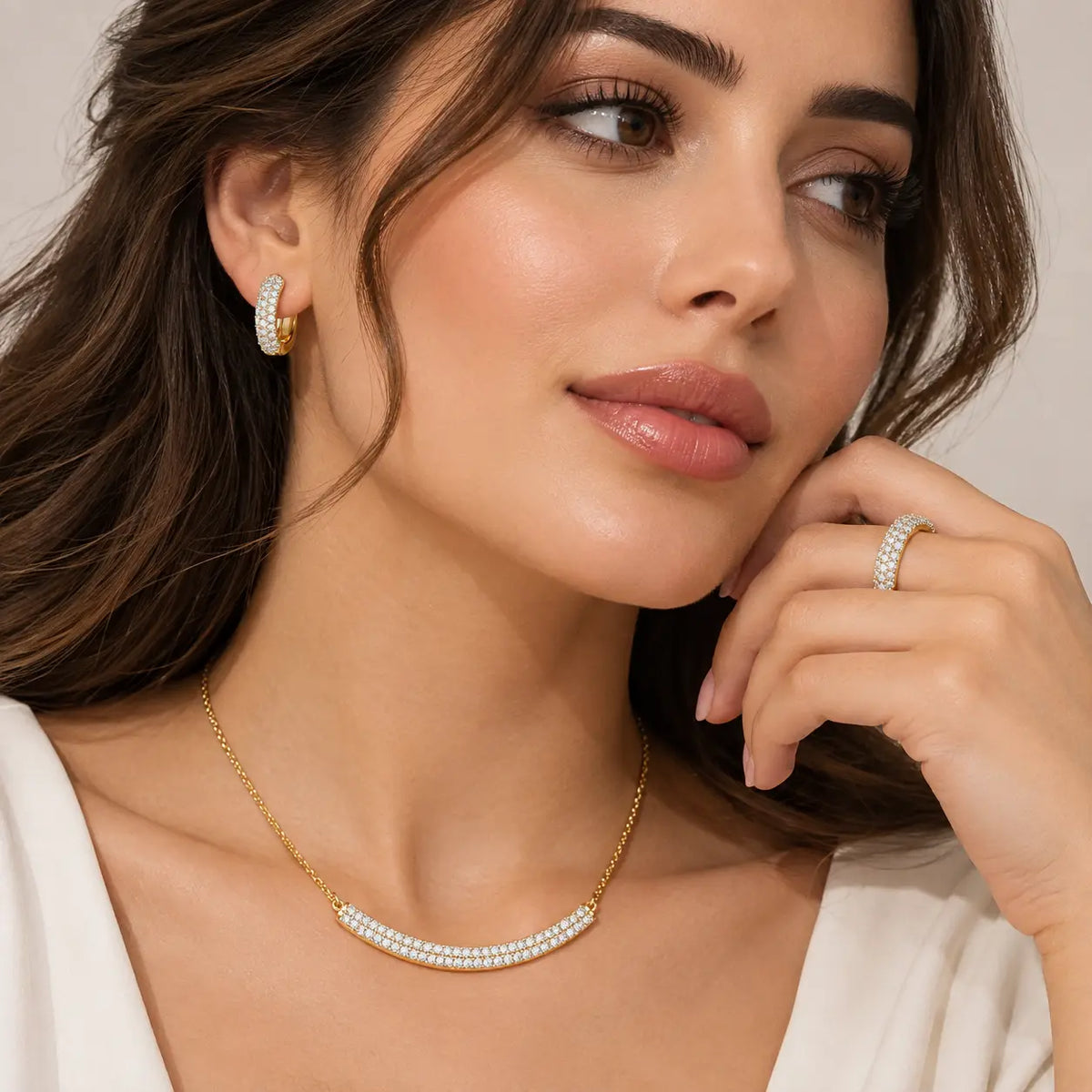 A close-up lifestyle shot of a woman wearing a gold necklace with a curved row of white pearls, a matching pearl hoop earring, and a pearl ring on her finger, set against a soft beige background.