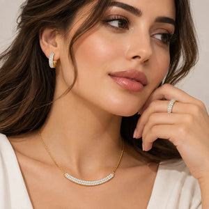 A close-up lifestyle shot of a woman wearing a gold necklace with a curved row of white pearls, a matching pearl hoop earring, and a pearl ring on her finger, set against a soft beige background.