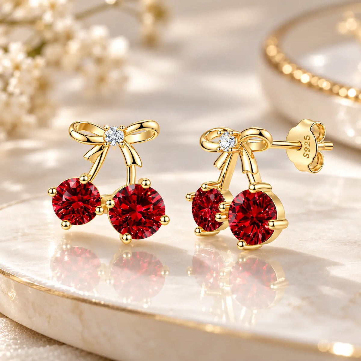 A pair of gold cherry stud earrings with deep red gemstone cherries, bow details, and a small clear center stone, displayed on a polished cream marble surface with soft floral props.