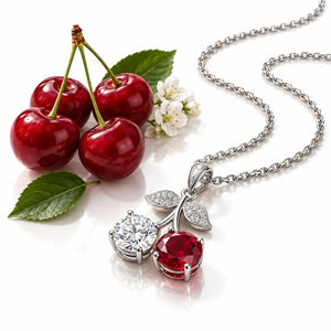 Silver-tone cherry pendant necklace on a white background, styled with red cherries, green leaves, and small white blossoms. The pendant features one clear round gemstone and one red round gemstone, with leaf-shaped pavé accents.