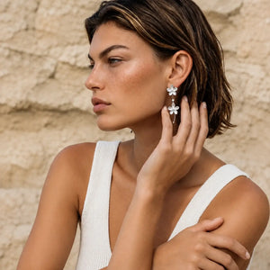 A woman in a white ribbed tank top poses against a textured beige stone wall, wearing a pair of silver dangle earrings with white flower charms.