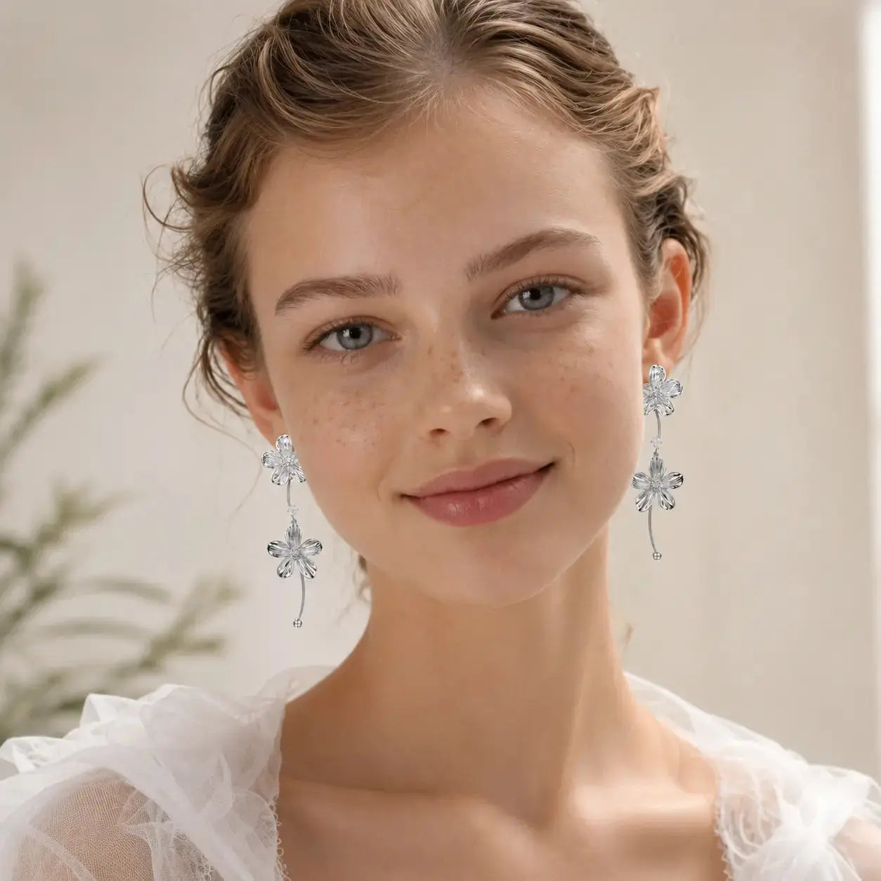 A close-up portrait of a woman wearing long, dangling silver-toned earrings with floral and vine-like details, against a soft light beige background.