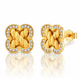 A pair of gold post earrings with a four-leaf clover shape, featuring a braided knot design in the center and a border of small clear stones, shown on a white background with a reflection.