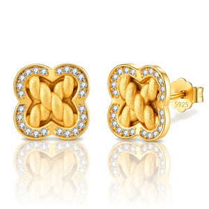 A pair of gold post earrings with a four-leaf clover shape, featuring a braided knot design in the center and a border of small clear stones, shown on a white background with a reflection.