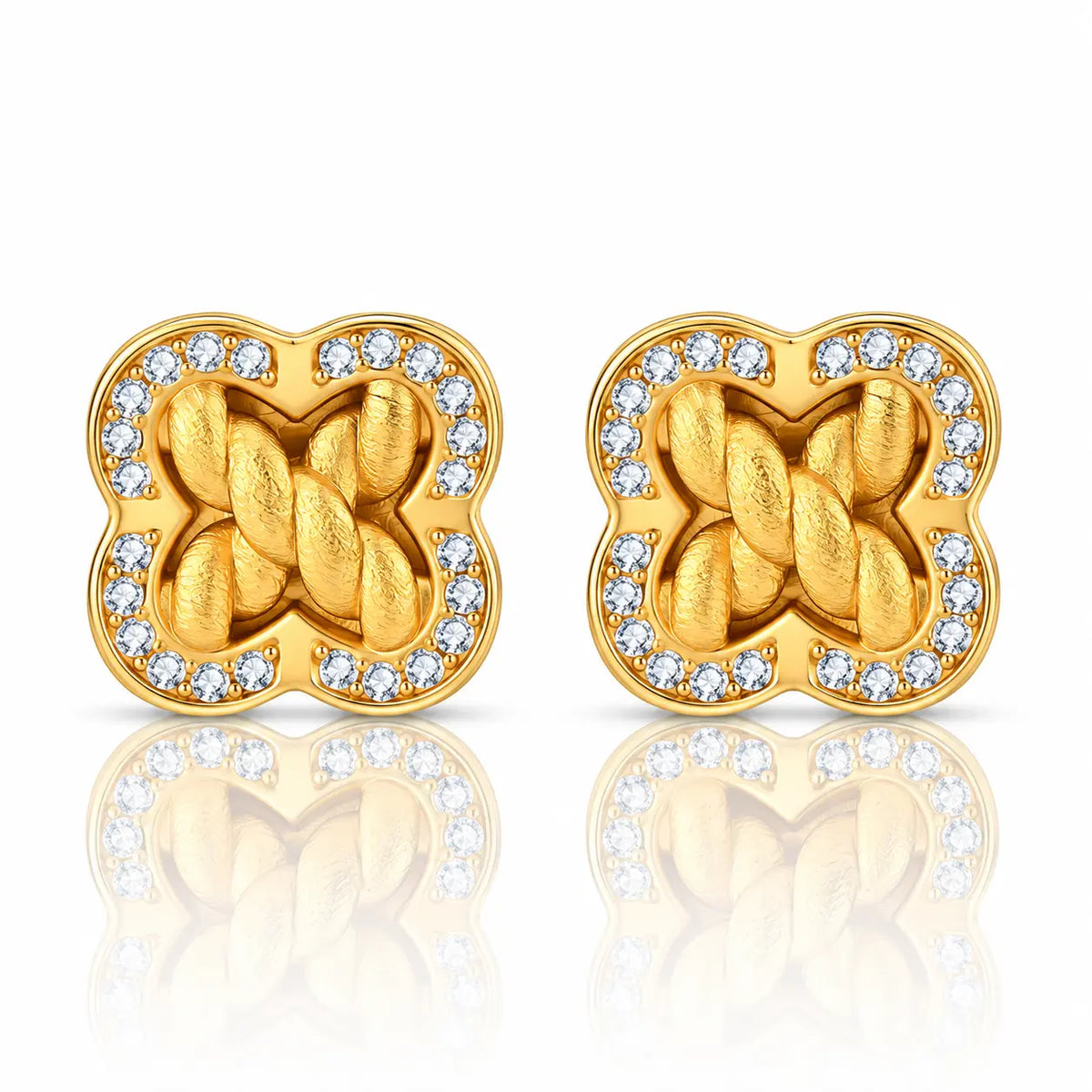 A pair of gold-toned stud earrings with a four-leaf clover shape, featuring a twisted knot design in the center and a border of small clear stones, shown on a white background with a soft reflection below.