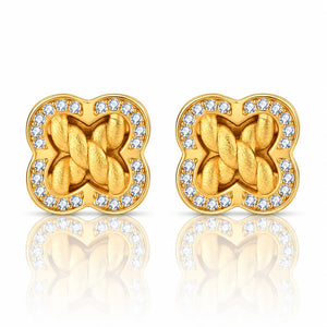A pair of gold-toned stud earrings with a four-leaf clover shape, featuring a twisted knot design in the center and a border of small clear stones, shown on a white background with a soft reflection below.