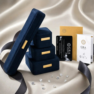 A luxury jewelry presentation set featuring three stacked navy blue velvet ring boxes with gold-tone accents, a single open box leaning against the stack, a gold envelope, branded black and white cards, and scattered clear gemstones on a draped champagne satin background.