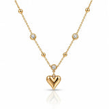 A gold necklace featuring a polished heart-shaped pendant and several small bezel-set clear stones along the chain, displayed on a clean white background with a soft reflection.