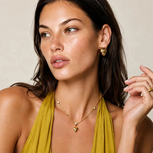 A close-up lifestyle shot of a model wearing a delicate gold necklace with a small heart pendant and matching gold hoop earrings, paired with a mustard yellow halter top against a neutral beige background.