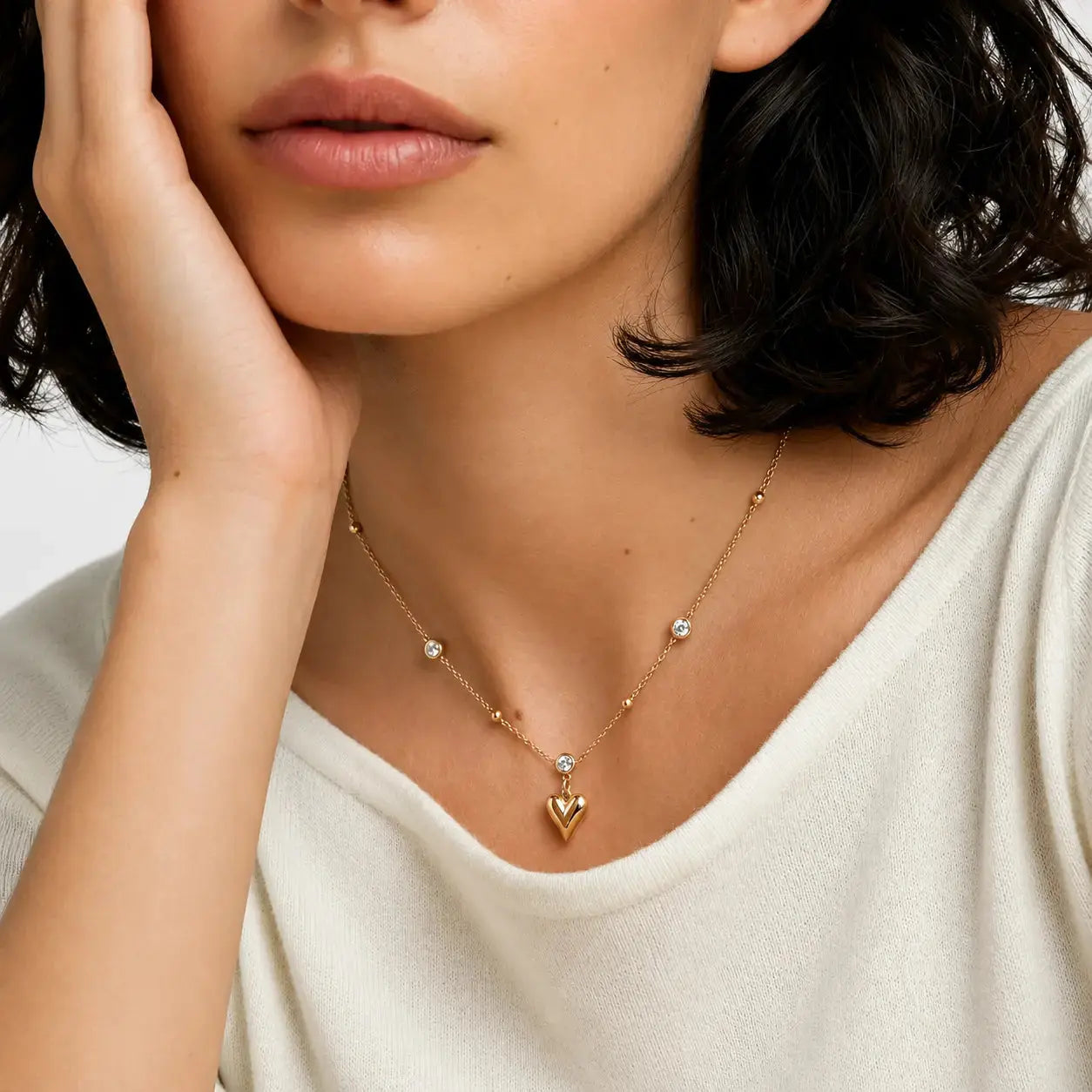 A close-up of a model wearing a delicate gold necklace with a small heart pendant and tiny clear stones, paired with a cream-colored top.