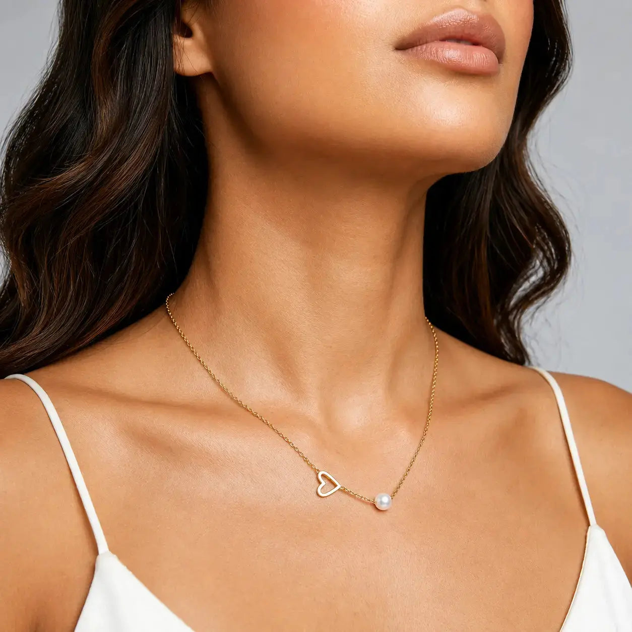 A close-up of a woman wearing a delicate gold chain necklace with an open heart and a small white pearl, styled with a white spaghetti strap top against a neutral gray background.