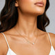A close-up of a woman wearing a delicate gold chain necklace with an open heart and a small white pearl, styled with a white spaghetti strap top against a neutral gray background.