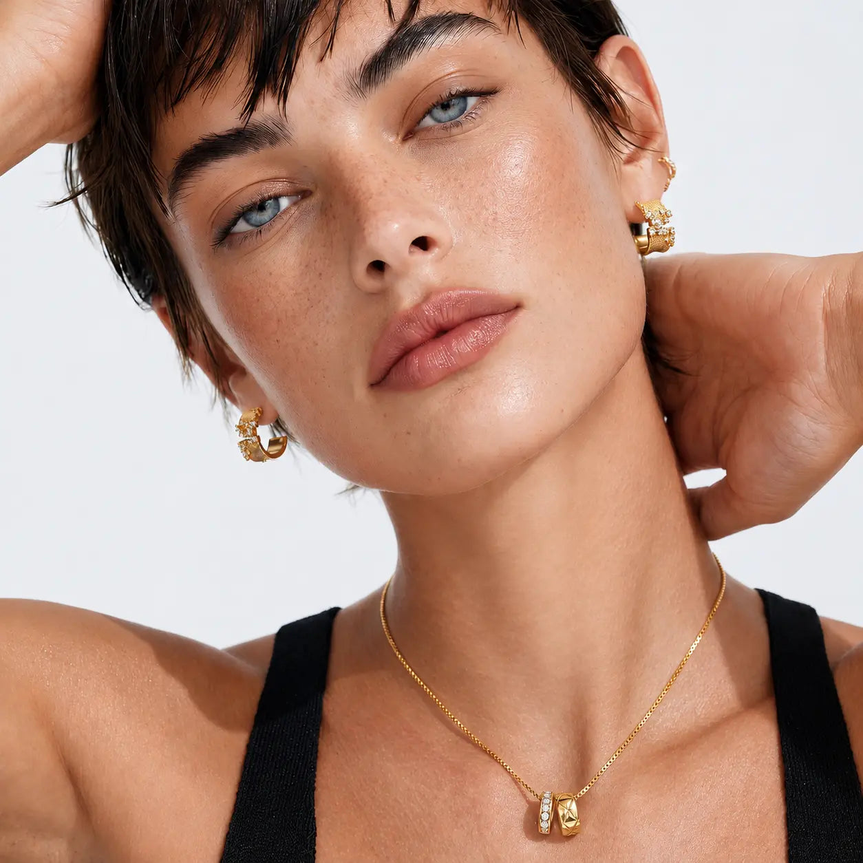 Close-up of a model wearing gold jewelry, including a delicate gold chain necklace with a small gold pendant, gold hoop earrings, and a gold headband, against a clean white background.