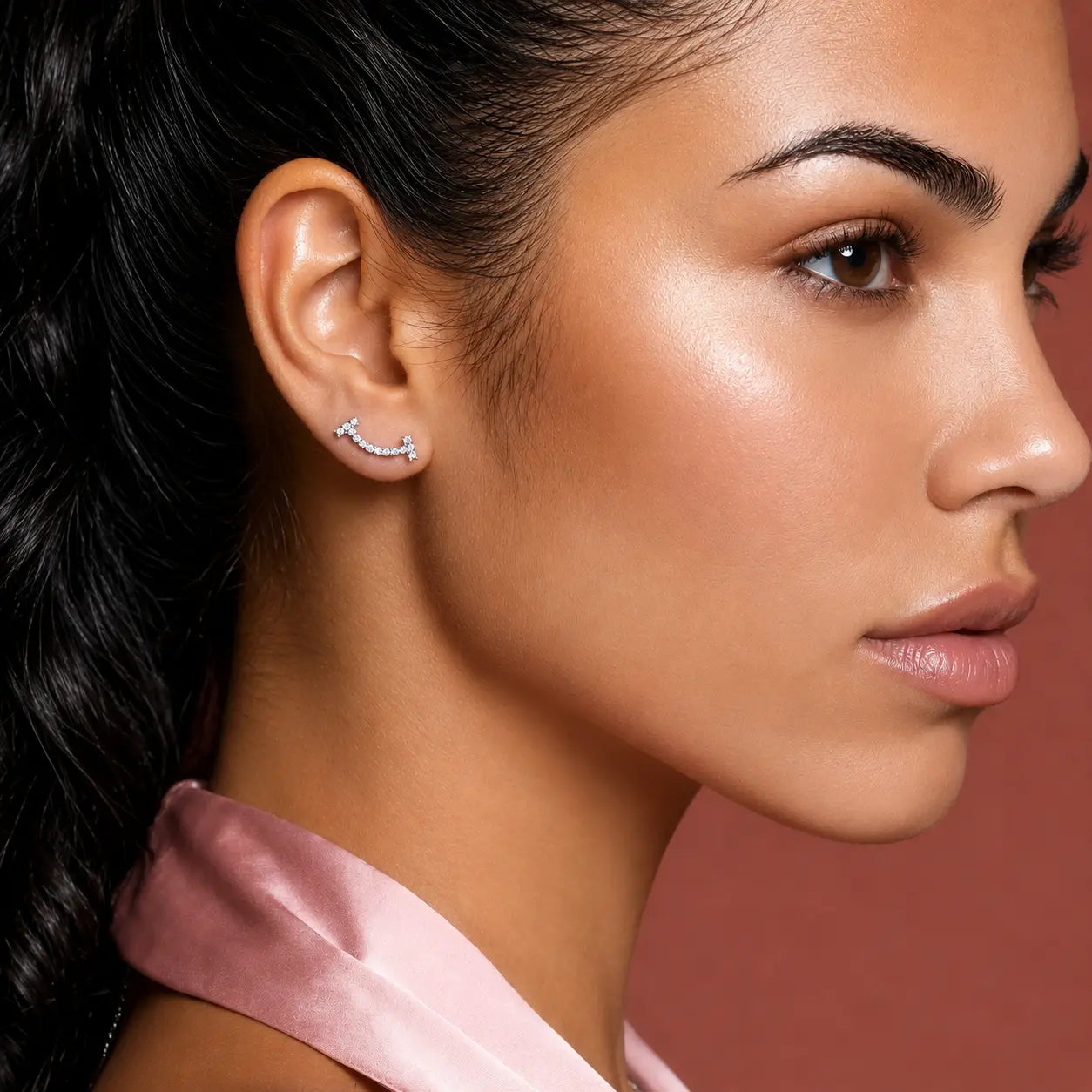 A close-up side profile of a woman wearing a small silver crescent-shaped stud earring with clear stones, set against a solid dusty rose background.