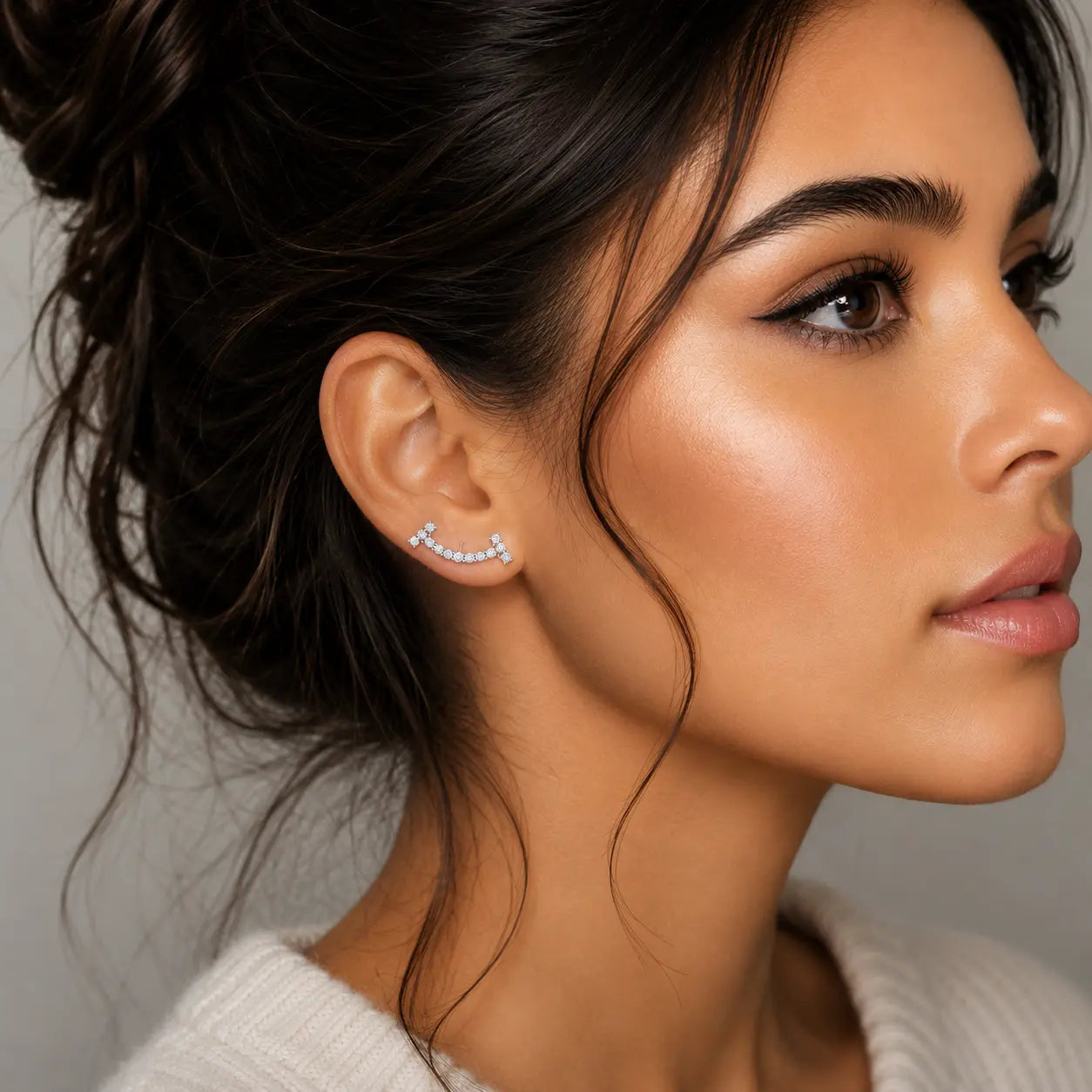 A close-up side profile of a woman with dark hair in an updo wearing a small silver crescent-shaped stud earring set with clear stones, against a soft gray background.