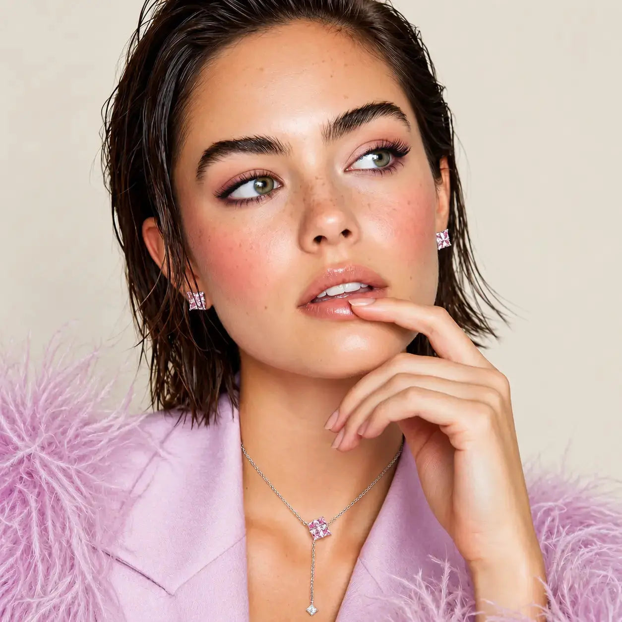 A close-up portrait of a woman wearing a delicate silver necklace with a pink gemstone and matching stud earrings, styled with a light pink feathered jacket against a soft beige background.