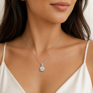 A close-up of a woman wearing a delicate silver chain necklace with a clear, flower-shaped gemstone pendant. She is wearing a white satin camisole against a soft, light background.