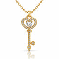 A gold key-shaped pendant necklace adorned with clear pavé stones and a heart-shaped clear center stone, suspended from a gold chain on a white background with a soft reflection below.