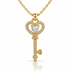 A gold key-shaped pendant necklace adorned with clear pavé stones and a heart-shaped clear center stone, suspended from a gold chain on a white background with a soft reflection below.