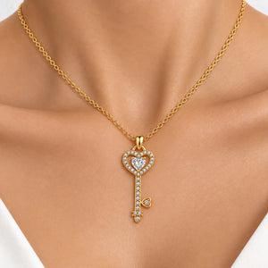 A gold key-shaped pendant necklace with a heart outline and clear stones, worn on a model's neck.