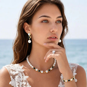 A woman posing with a pearl and gold jewelry set against a soft-focus ocean background. The set includes a pearl necklace with gold and black bezel-set round stones, matching drop earrings, a ring, and a bracelet.