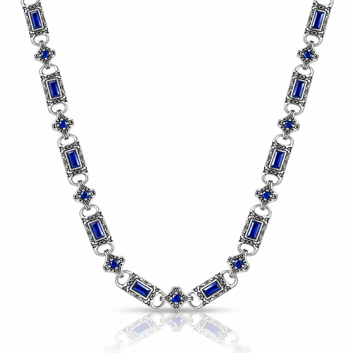 A silver-toned link necklace featuring rectangular blue gemstones and small blue floral accents, displayed on a clean white background with a soft reflection below.
